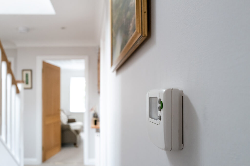 White hallway with a digital thermostat on the wall. A wooden staircase and an open door leading to a bright room create a calm and inviting atmosphere.
