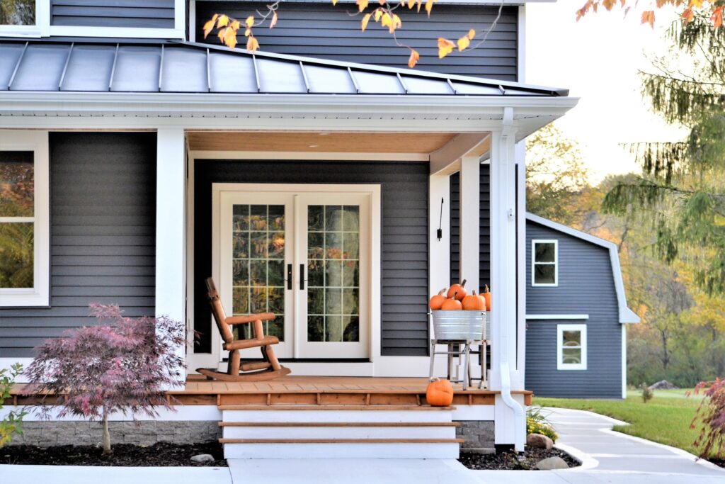 A cozy porch with a wooden rocking chair beside some fall decor. Autumn leaves and warm light enhance the welcoming, seasonal atmosphere.
