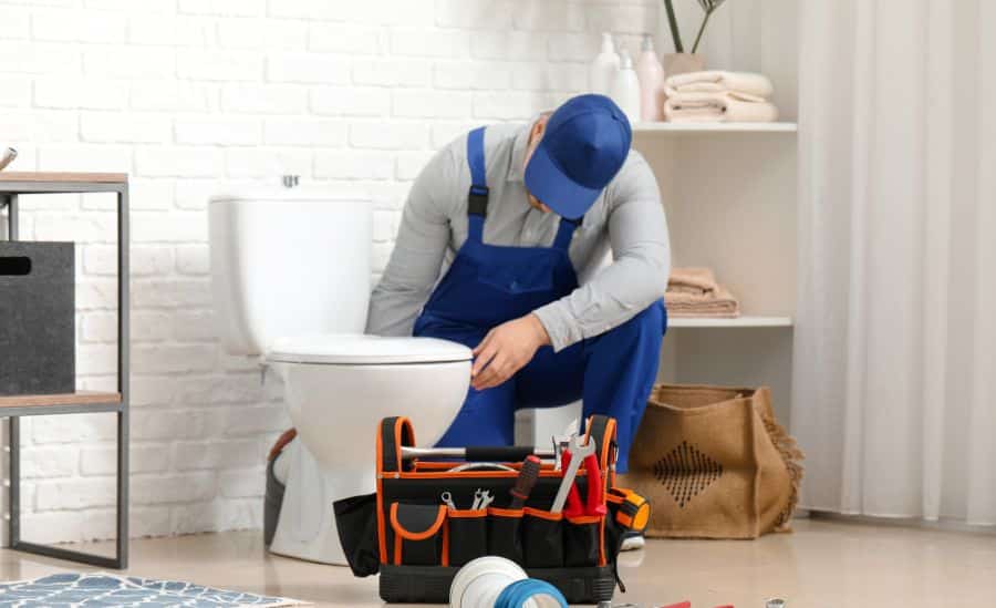 A plumber in blue overalls and cap examines a toilet in a bathroom, with a tool bag and tools on the floor beside him.