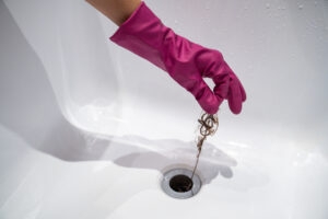 A hand in a pink rubber cleaning glove is lifting hair out of the clogged drain of the bathtub, illustrating the importance of a drain clear.