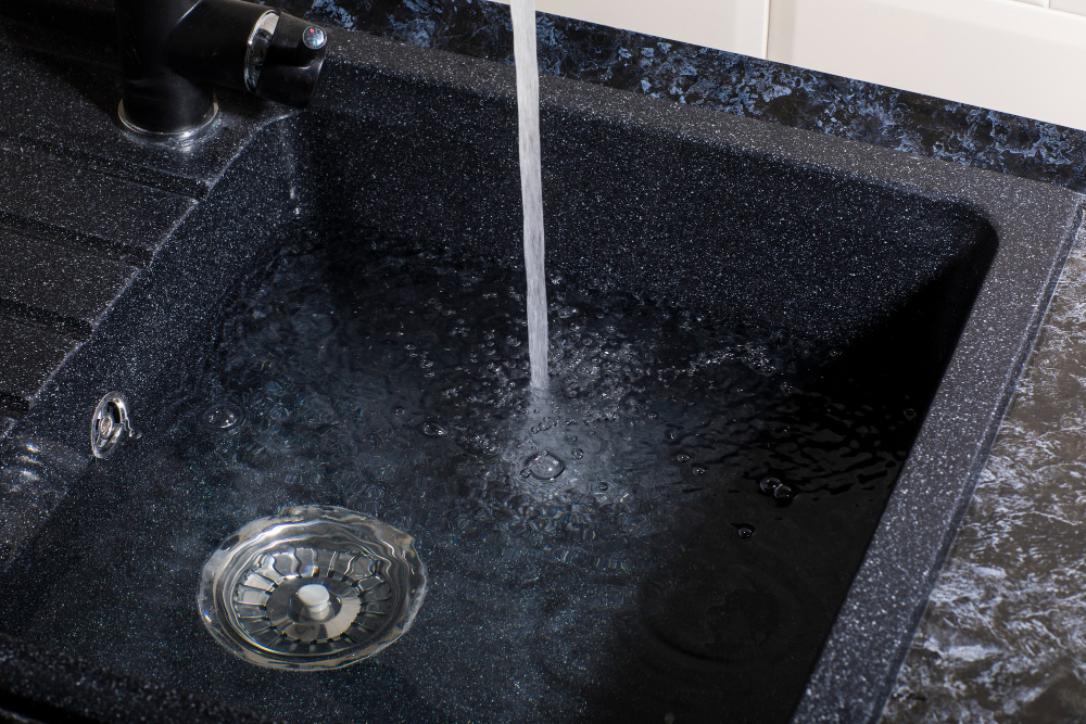 A kitchen sink has water filling it up. The drain is plugged by a drain cover to prevent the water from escaping.