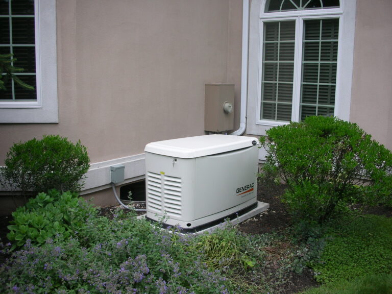 A standby generator sits on a concrete pad outside a house, surrounded by bushes and plants near a window with closed blinds.