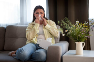 Woman sneezing on a couch, surrounded by allergens possibly missed by a clogged or low-rated MERV air filter.