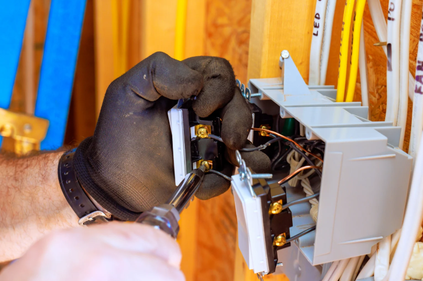 A person wearing black gloves uses a screwdriver to install electrical outlets in a junction box with exposed wiring inside a wooden wall frame.