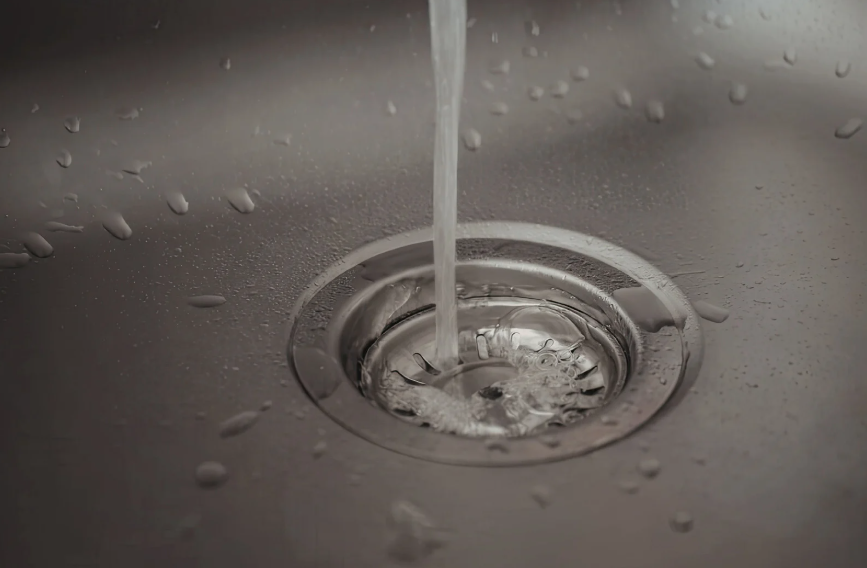 Water flows from a faucet into a stainless steel sink, forming small bubbles and droplets around the drain.