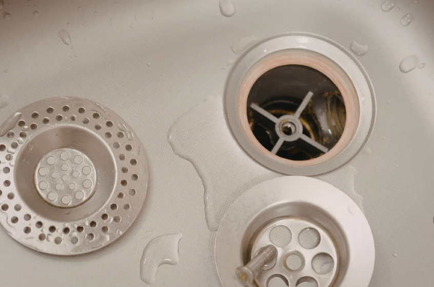 Close-up of a stainless steel sink with a strainer and stopper removed, exposing the drain opening and some water droplets on the surface.