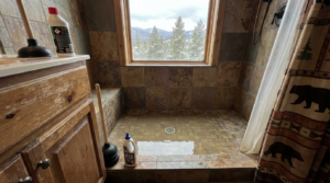 A tiled shower with standing water, cleaning supplies, and a plunger, next to a window showing a snowy mountain landscape.