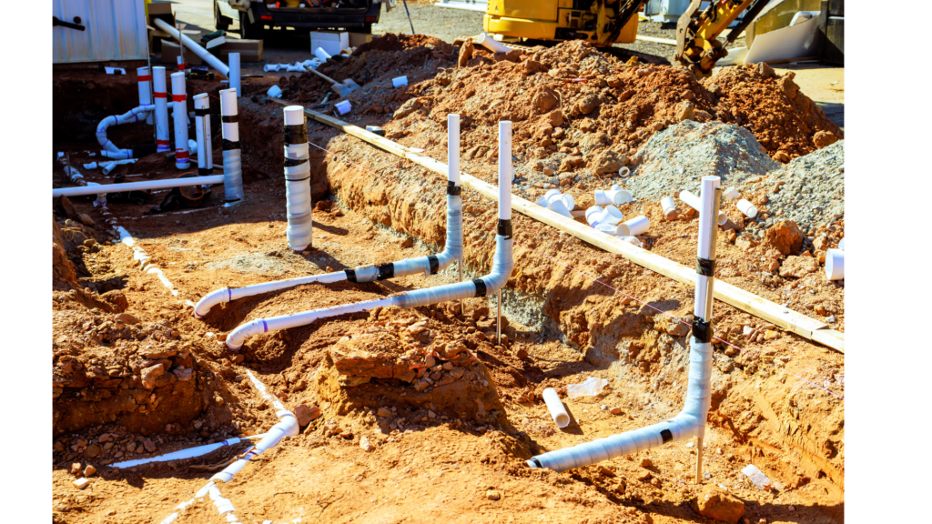 PVC plumbing pipes and fittings are exposed in a trench at a construction site, with soil piles and construction equipment visible in the background.