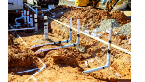 PVC plumbing pipes and fittings are exposed in a trench at a construction site, with soil piles and construction equipment visible in the background.