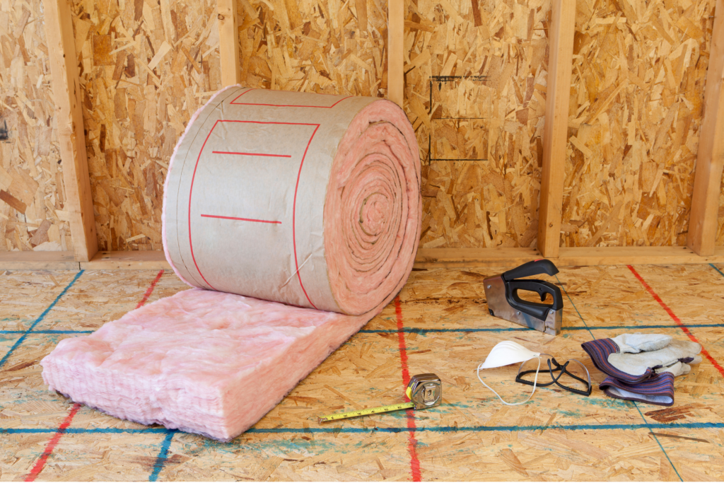 A roll of pink insulation, gloves, face mask, tape measure, utility knife, and stapler are on a plywood floor against a wooden frame wall.
