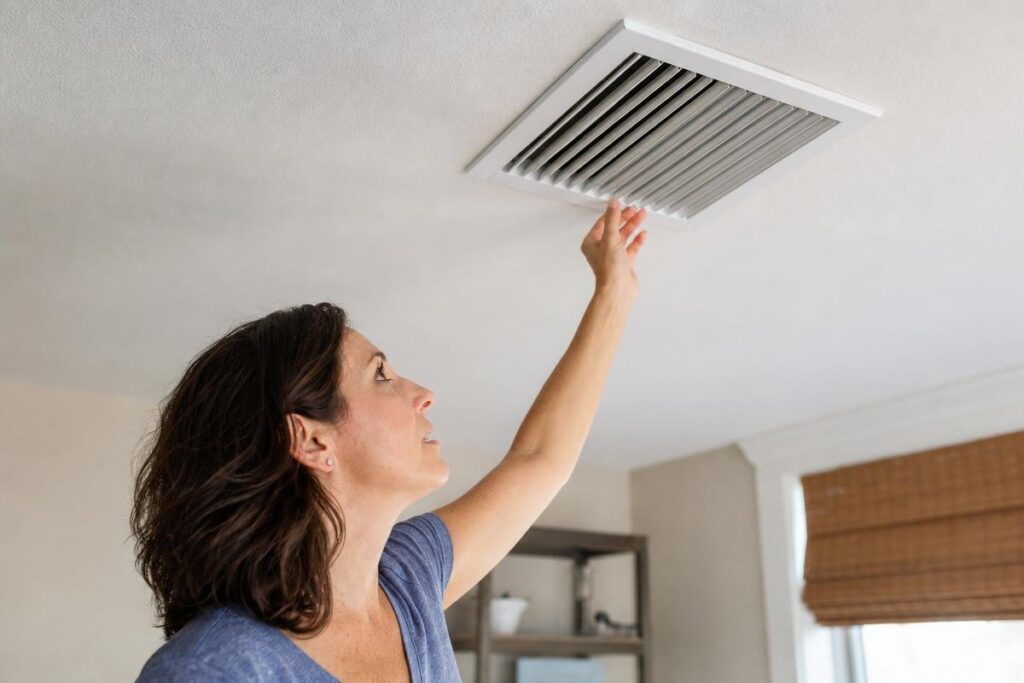 A woman reaches up to adjust a ceiling air vent in a room with white walls and a window covered by a bamboo shade.