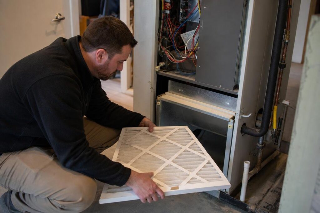 A man kneels and installs a new air filter into an open furnace unit inside a residential space.