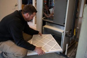 %Focuskw% | Precision Plumbing &Amp; Heating A man kneels and installs a new air filter into an open furnace unit inside a residential space.