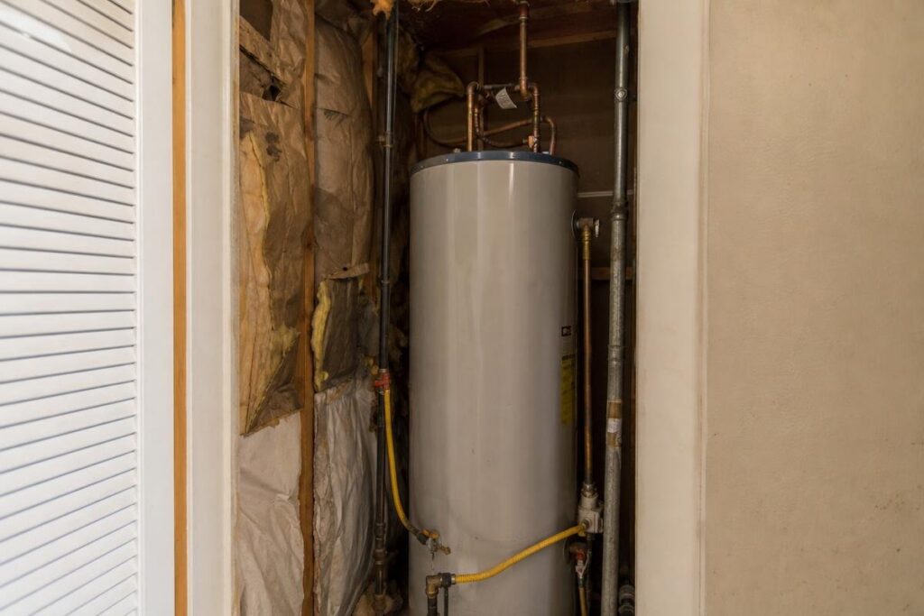A water heater is installed in a narrow utility closet with exposed insulation and pipes visible on the surrounding walls.
