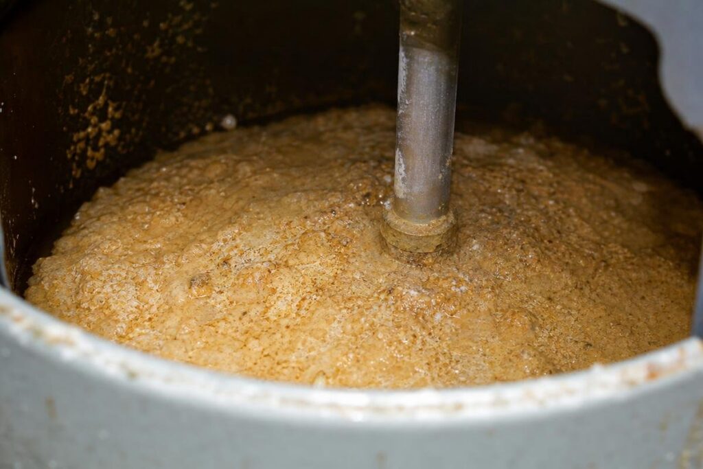 Thick, bubbly brown mixture being stirred by a metal rod inside a large industrial container.