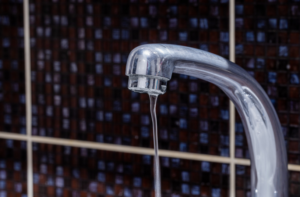 %Focuskw% | Precision Plumbing &Amp; Heating A close-up of a chrome faucet with a thin stream of water running, set against a background of dark brown and beige tiled wall.