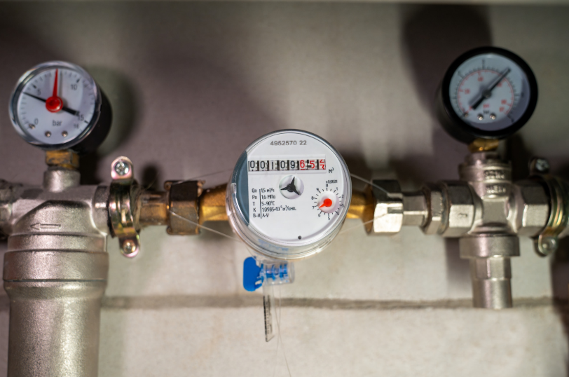 Three metal pipes with attached pressure gauges and a central water meter mounted on a wall, displaying readings and calibration markings.