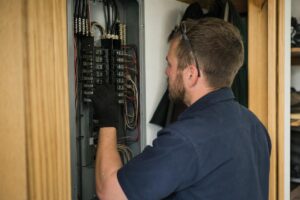 A man wearing gloves and safety glasses adjusts switches inside an electrical breaker panel mounted on a wall.