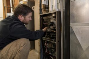 A man inspects and repairs the internal components of a furnace unit in a basement or utility room.