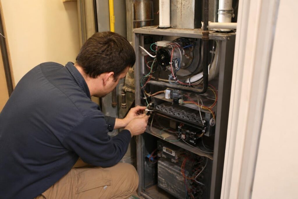 A person kneels in front of an open furnace, using a tool to adjust or repair wiring and components inside the unit.
