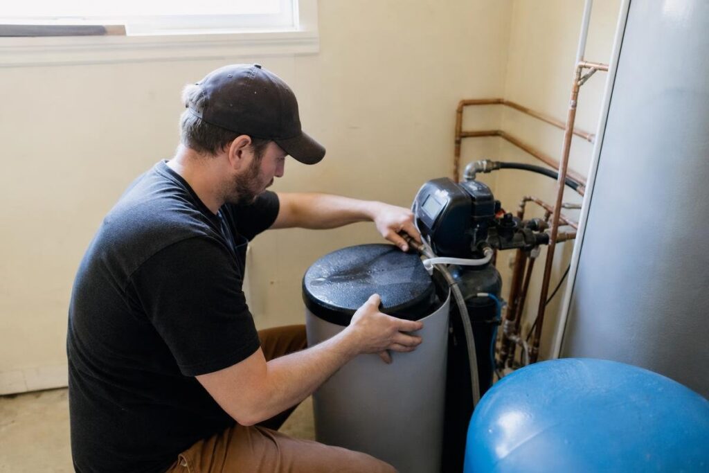 A man adjusts the lid of a water softener system in a utility room, with pipes and a pressure tank visible nearby.