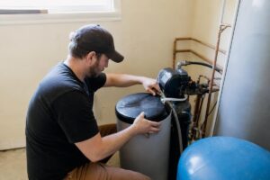 A man adjusts the lid of a water softener system in a utility room, with pipes and a pressure tank visible nearby.